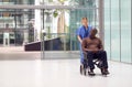 Female Nurse Wearing Scrubs Wheeling Patient In Wheelchair Through Lobby Of Modern Hospital Building Royalty Free Stock Photo