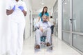 Female Nurse Wearing Scrubs Wheeling Patient In Wheelchair Through Lobby Of Modern Hospital Building. Doctors and nurse in Royalty Free Stock Photo