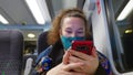 Female passenger wearing face covering mask during covid-19 lockdown using phone inside train in england uk