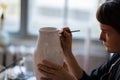 Focused brunette woman paints text with brush on white ceramic vase at table on blurred background