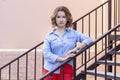 Female portrait. beautiful young girl stands on the stairs with iron railing. red pants and a denim shirt.