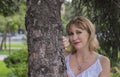 Charming pretty middle-aged woman with blond hair, 50-55 years old, standing, smiling, leaning against a tree Royalty Free Stock Photo