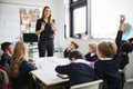 Female primary school teacher standing in a classroom gesturing to schoolchildren sitting at a table listening, one raising their Royalty Free Stock Photo