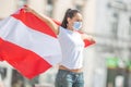 Female in protective mask holds a flag of Austria behind her outdoors on a sunny day