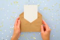 Female`s hands holding kraft paper envelope letter with blank white card mockup over pastel blue background with golden confetti Royalty Free Stock Photo