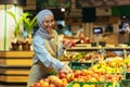 Female seller in hijab browsing and checking apples in supermarket, woman in apron smiling at work in store in fruit and