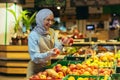 Female seller in hijab browsing and checking apples in supermarket, woman in apron smiling at work in store in fruit and