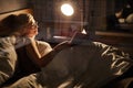 female in spectacles lying on bed reading favourite interesting book at home in room lighted by lamp