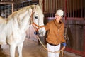 Female stable worker leads a white horse out of stall