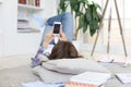 Female student checking social media before getting back to studying, lying on floor against cozy domestic interior