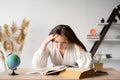 female student with headache and fatigue sits at the table and studies mathematics with textbook and notebook. An