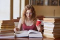 Female student posing with coffee in hand, suraunded with stacks of books at reding room. Young woman sitting at table doing