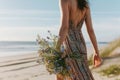 female in a sundress holding wildflowers, walking on a beach