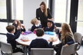 Female teacher stands talking to a group of primary school kids sitting together at a round table eating their packed lunches