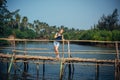 A female tourist in denim shorts with short haircut on sunny tropical day. Young pretty girl stands on wooden bridge over small Royalty Free Stock Photo