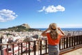 Female tourist holding hat and looking at Alicante cityscape and Mount Benacantil with Santa Barbara Castle and sea on background