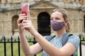 Female Tourist On Vacation Wearing Face Mask During Covid-19 Pandemic Taking Photo On Mobile Phone In Oxford UK