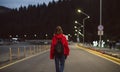 Female traveler in a red raincoat walking down the road in the mountainous scenery in the evening. Young tourist girl back view on