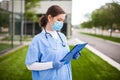 Female UK NHS doctor holding blue clipboard standing outside hospital clinic street entrance Royalty Free Stock Photo