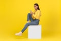 Female Using Smartphone Texting Sitting On Cube Over Yellow Background