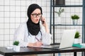 Female veiled scientist sitting on her desk looking at laptop opens her mouth with shocked face emotion while talking on