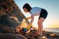 A female volunteer collects plastic bottles in a garbage bag. In the background, the sea and the sunset. The concept of Royalty Free Stock Photo