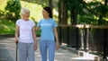 Female volunteer and senior patient with stick walking hospital garden, care