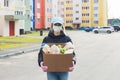 Female volunteer wearing a protective mask walks down the street with a box of groceries, donations