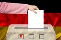 Female voter drops a ballot in a transparent ballot box against the background of the national flag of Germany, concept of state