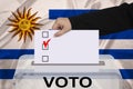 Female voter drops a ballot in a transparent ballot box on the background of the national flag of Uruguay, concept of state