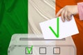 Female voter drops a ballot in a transparent ballot box against the background of the national flag of Ireland, concept of state