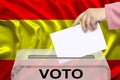 Female voter drops a ballot in a transparent ballot box against the background of the national flag of Spain, concept of state Royalty Free Stock Photo