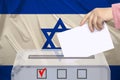 Female voter lowers the ballot in a transparent ballot box against the background of the national flag of Israel, concept of state