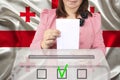 Female voter lowers the ballot in a transparent ballot box on the background of the national flag of Georgia, concept of state