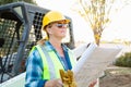 Female Worker Holding Technical Blueprints Near Small Bulldozer