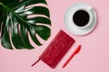 Female workspace with red notebook and pen, coffee cup and green monstera leaf on pink background, top view Royalty Free Stock Photo