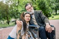 Female young social worker and disabled man on a walk. Girl and grandfather sitting in a wheelchair, hugging and posing