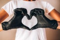 Females hands in black latex gloves show heart shape. Young slim tan woman in white tshirt and black gloves