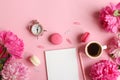 Feminine workplace with notepad, pencil, alarm clock, peony flowers, a cup of coffee and macarons on a pink background