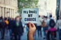 Feminist hands holding a protest banner with the message my body my choice over a crowded street. Human rights concept against