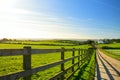 Fence casting shadows on a road leading to small house between scenic Cornish fields under blue sky, Cornwall, England