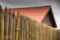 A fence made of sharp wooden stakes against the background of a wooden house with a red tiled roof