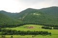 Fenced with a rectangular fence farm on the slope of a gentle hill, surrounded by a dense coniferous forest under a cloudy summer Royalty Free Stock Photo