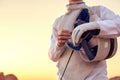 Fencer wearing white fencing costume and holding his fencing mask and a sword on sunny background