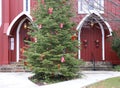 A soft gold light shines over the arched red church doors with colorful wreaths and a decorated tree dividing the pathway.