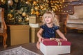 Festive little blonde girl in glitter dress opening a gift at home in the living room with Christmas decoration