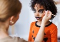 Festive makeup for Halloween. Woman doing pumpkin make-up for ethnic curly boy in costume while preparing holiday