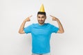 Festive man wearing T- shirt standing and pointing at yellow party cone, celebrating his birthday.