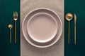 Festive place setting with beige napkin. Empty plates and gold cutlery on dark green background. Top view. Dining table in luxury