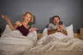 Lifestyle portrait of husband or boyfriend using mobile phone in bed with angry frustrated wife or girlfriend feeling ignored and